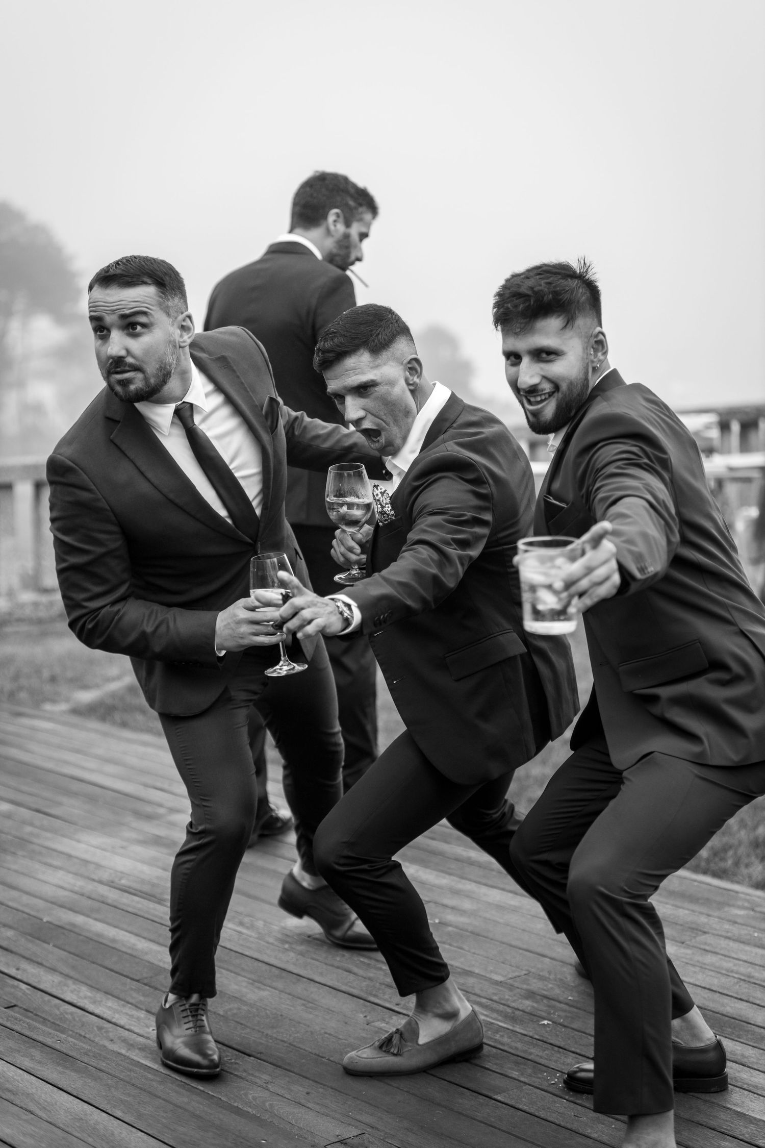 Three groomsmen in black suits strike a playful pose while holding drinks on a wooden deck.