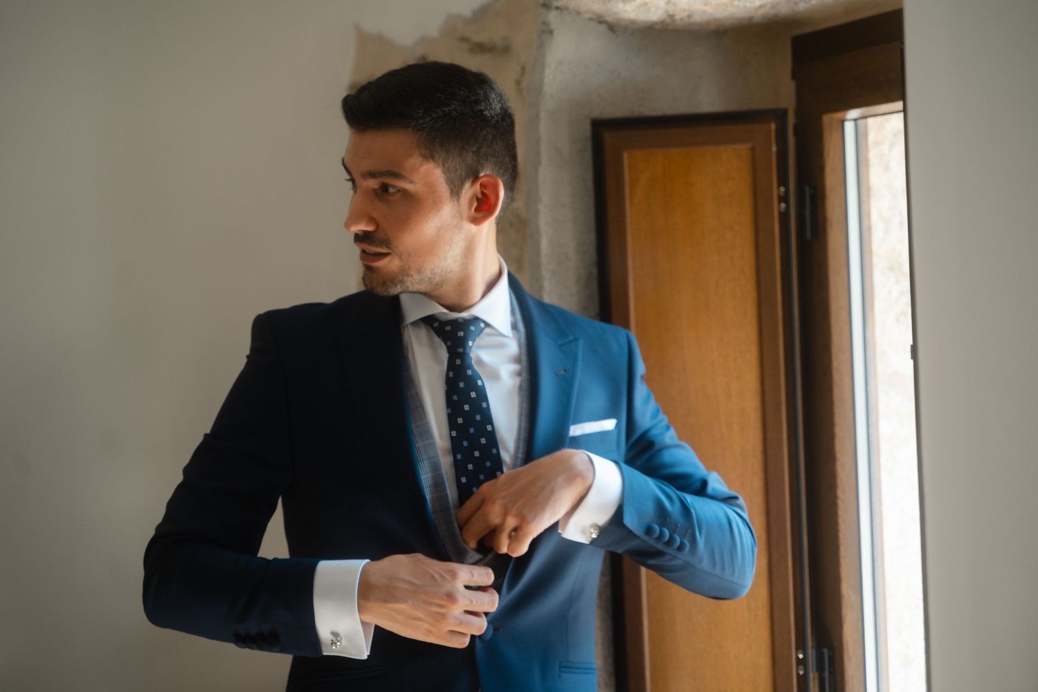 Man in navy suit and tie adjusting his jacket in a room with natural lighting.