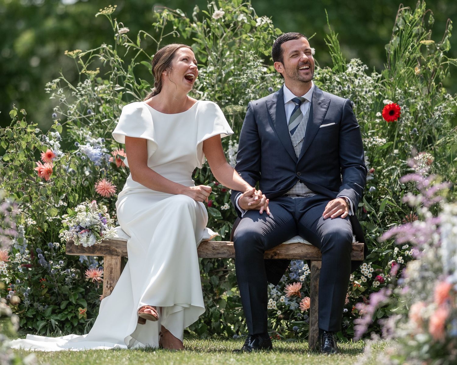 A couple in wedding attire shares a candid moment together in a lush garden setting with blooming flowers.
