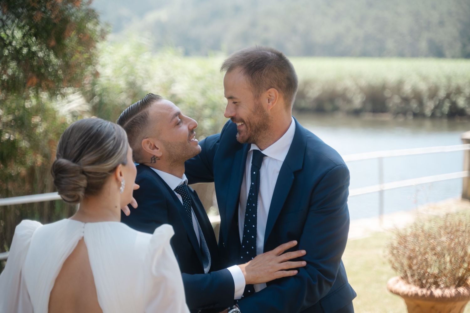 Three people in formal wedding attire share a joyful moment together by a waterfront on a sunny day.