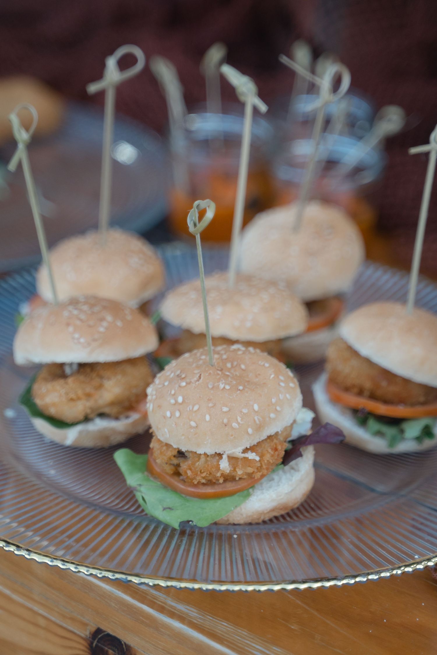 Mini sliders with sesame seed buns and lettuce served on a clear glass platter at a party.