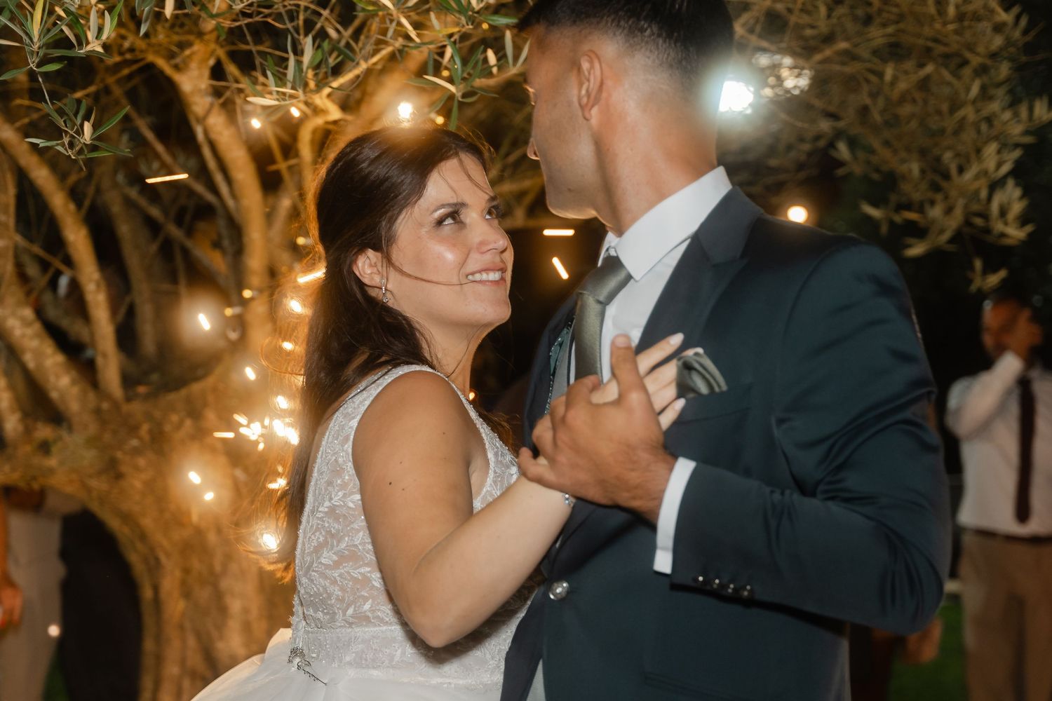 A couple shares a romantic dance under illuminated trees at an outdoor evening wedding reception.