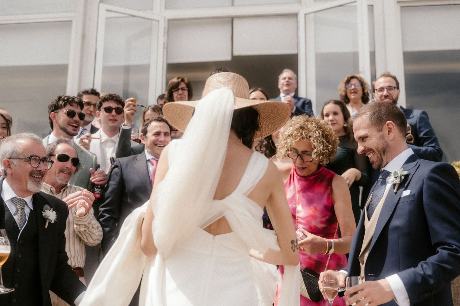 A group of wedding guests gather outdoors in formal attire, smiling and celebrating during a sunny daytime reception.