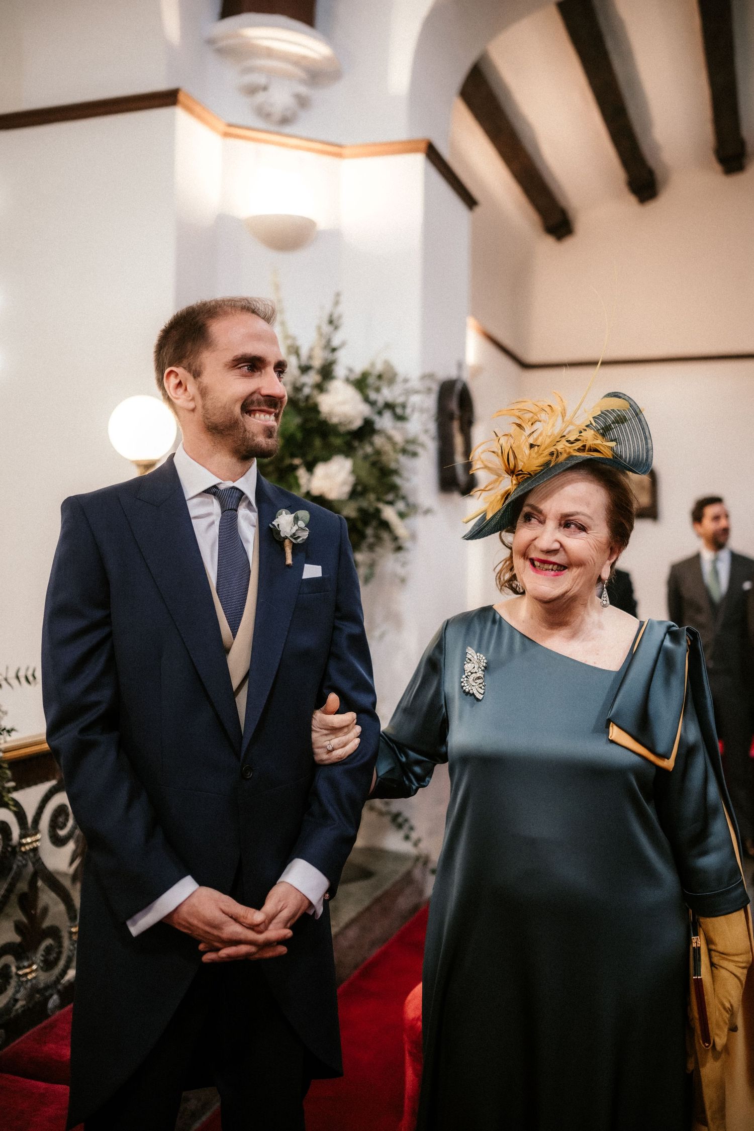Wedding guests share candid moments and laughter during an indoor reception with rustic wooden beams and warm lighting.