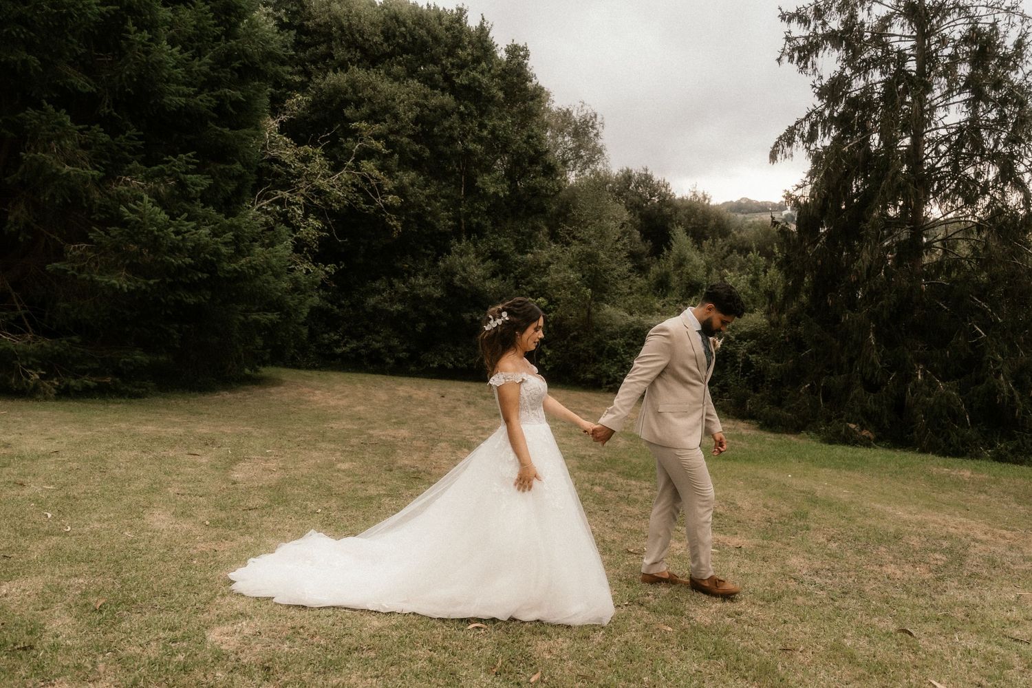 Romantic couple in wedding attire playfully runs and dances together across a grassy hillside during their outdoor celebration.