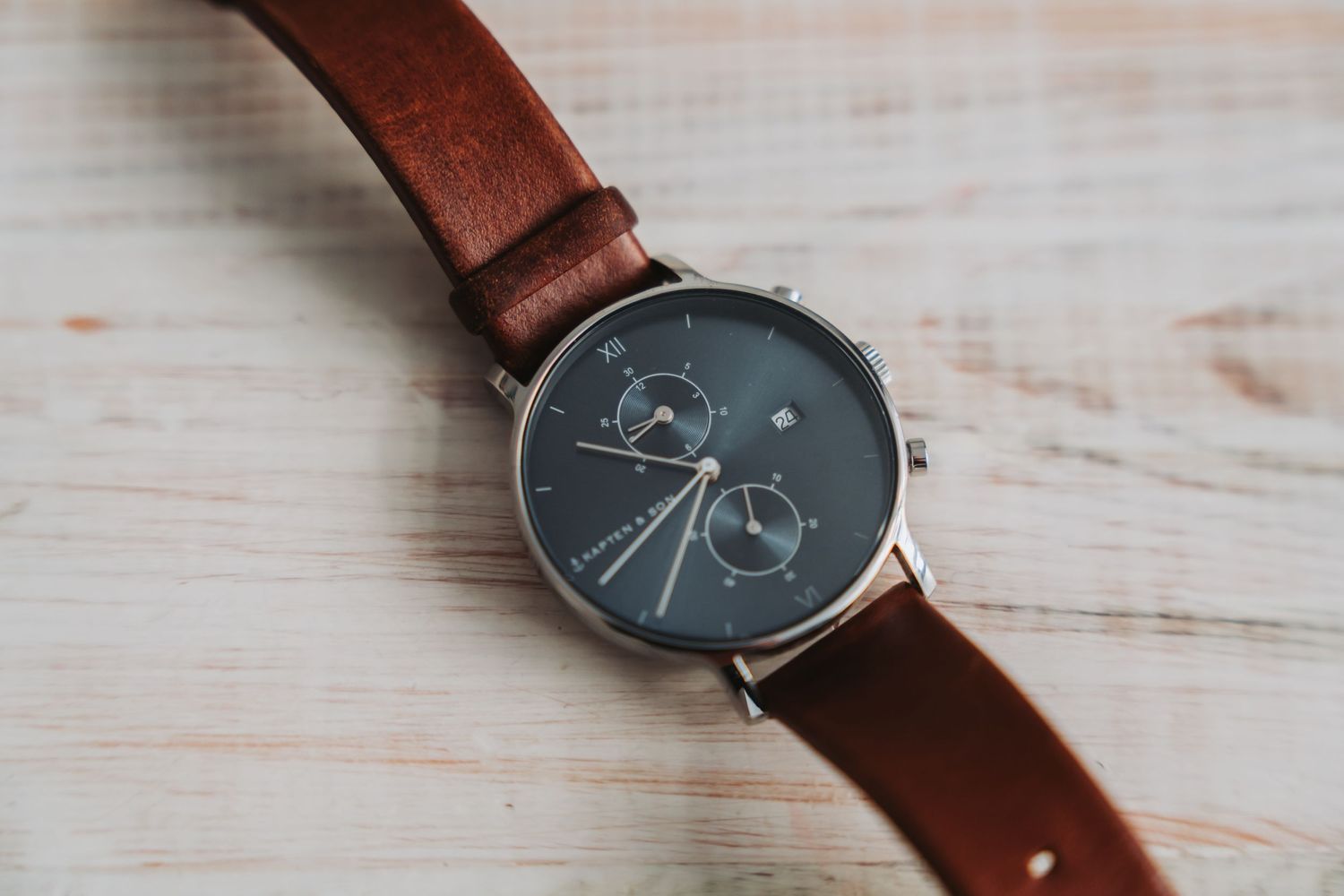 Brown leather watch strap with minimalist dark watch face displayed on white wooden surface.