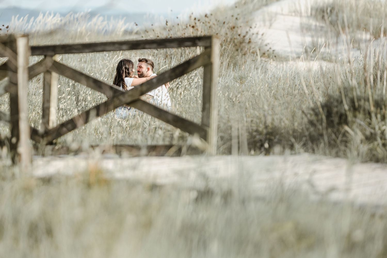 A couple embraces through a rustic wooden fence on a dreamy, sunlit day in nature.