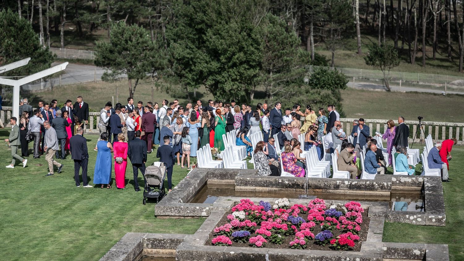 A large outdoor gathering of people in formal attire standing around a colorful garden bed during a special event.