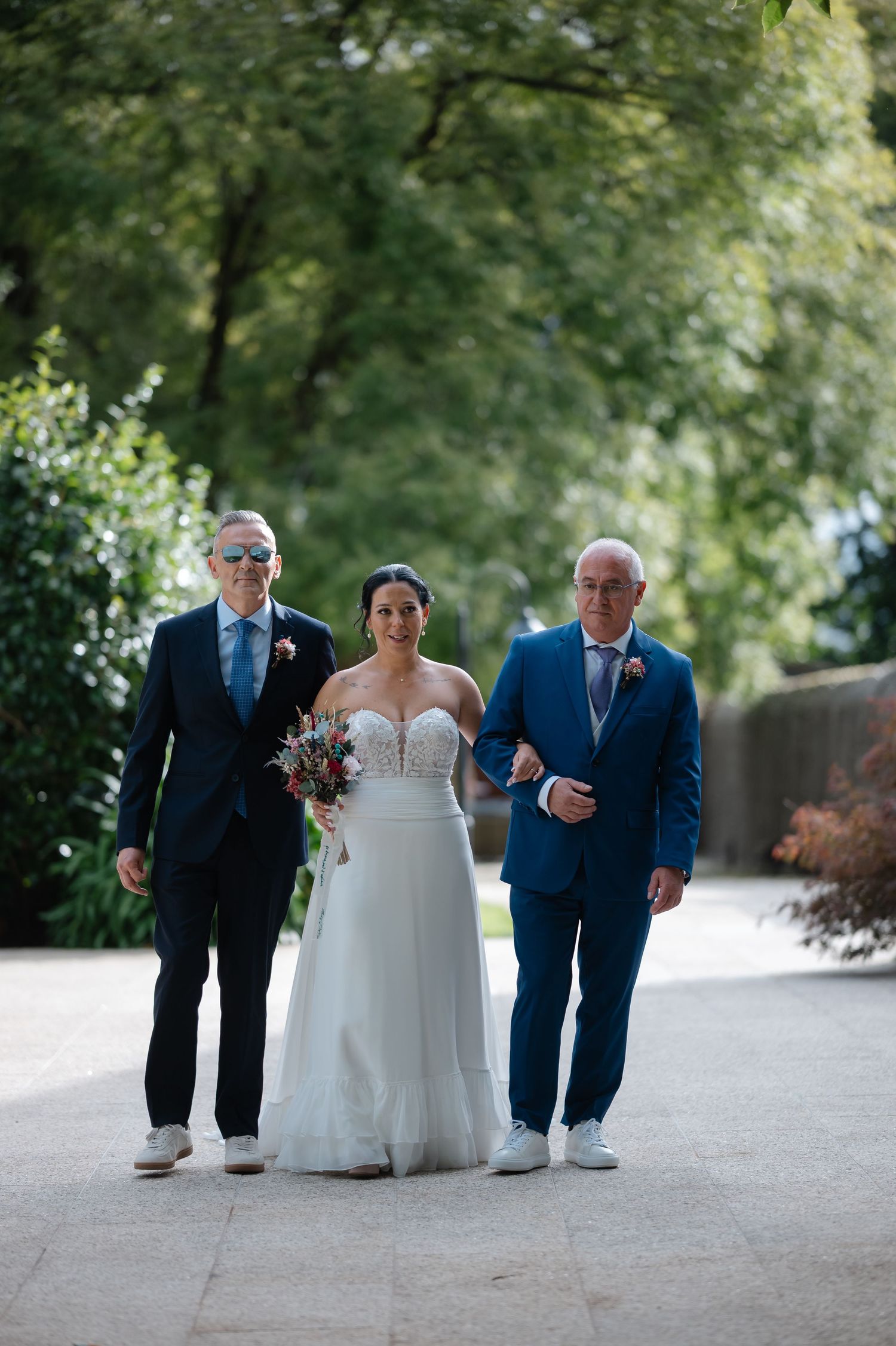 A wedding party walks together on a path surrounded by lush green trees and foliage.