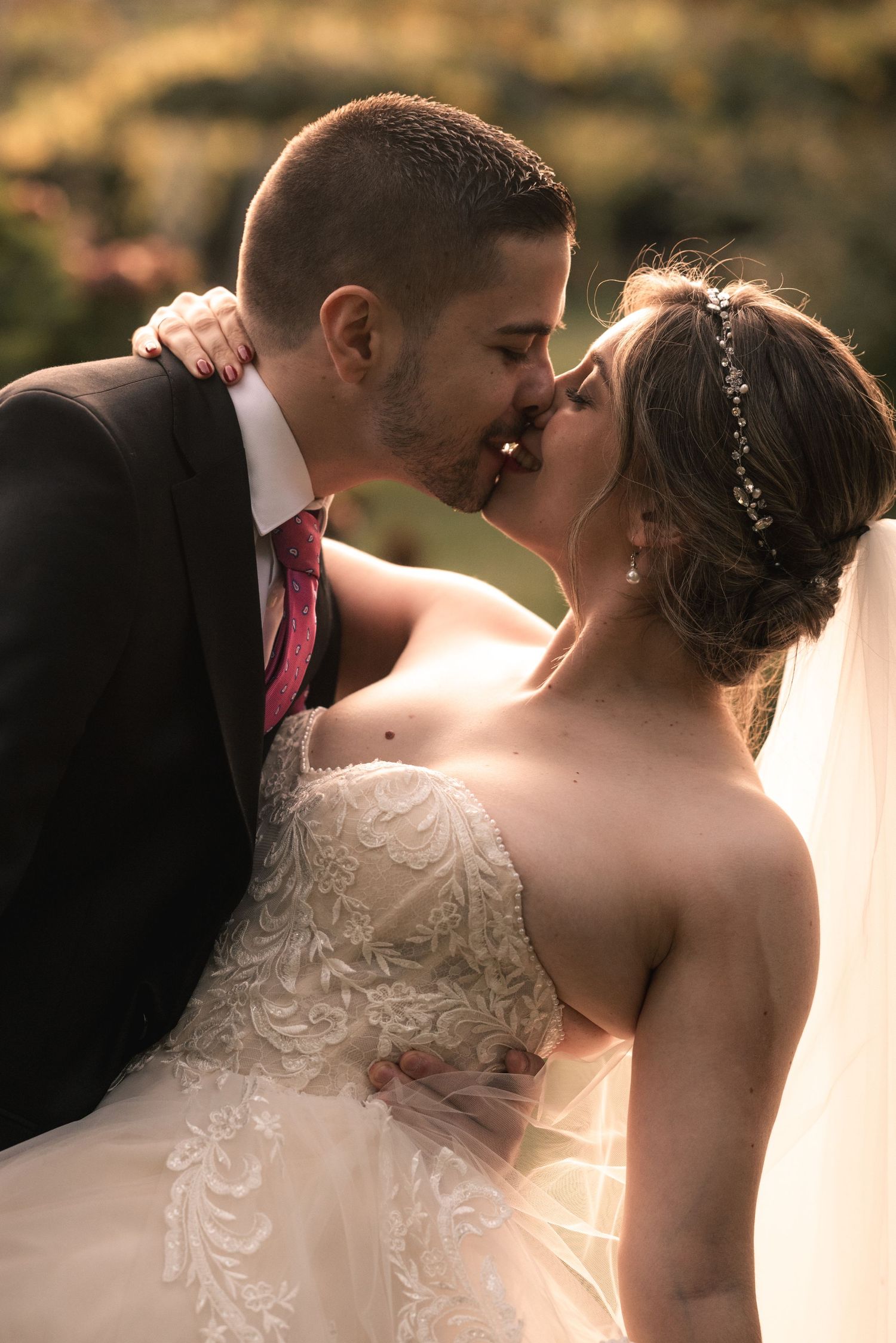 A romantic wedding couple shares a tender kiss outdoors with golden sunlight glowing behind them.