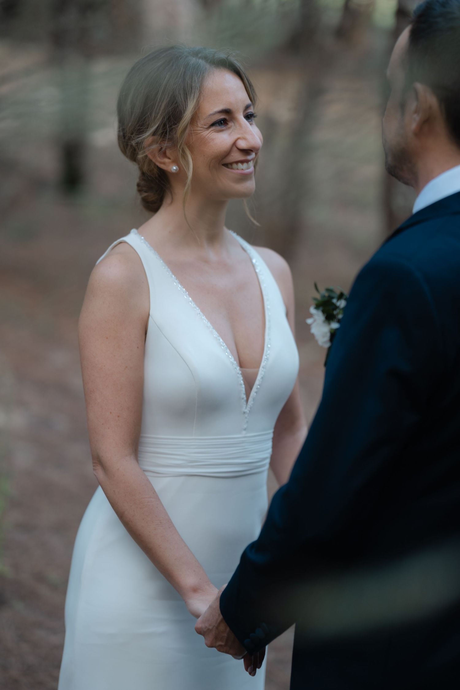 A bride in a sleek white dress shares a joyful moment with someone in formal wear during an outdoor wedding.