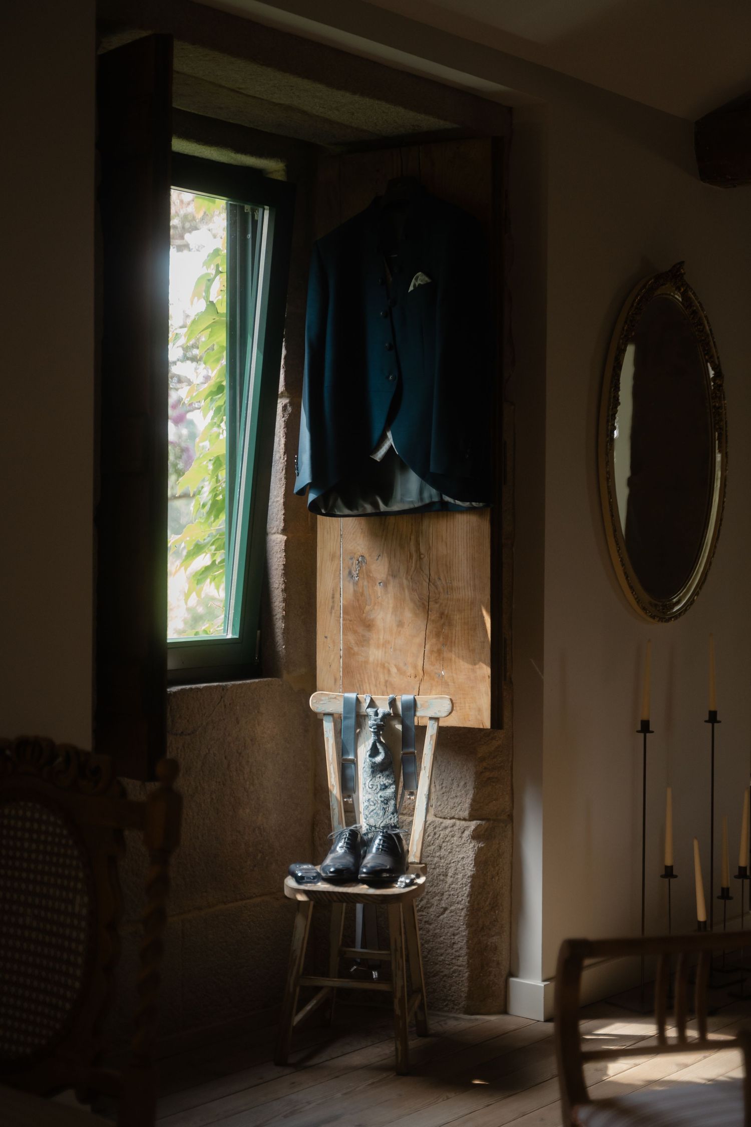 Moody interior scene with window light illuminating hanging clothes and vintage oval mirror on brick wall.