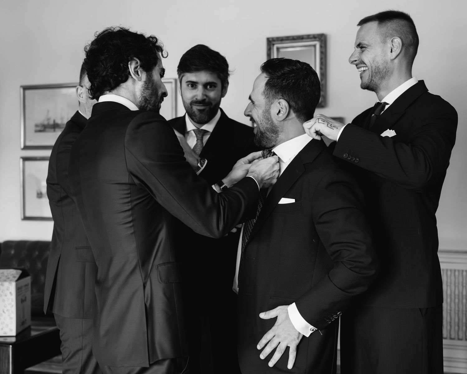 Groomsmen in black suits help each other get ready before a wedding ceremony.