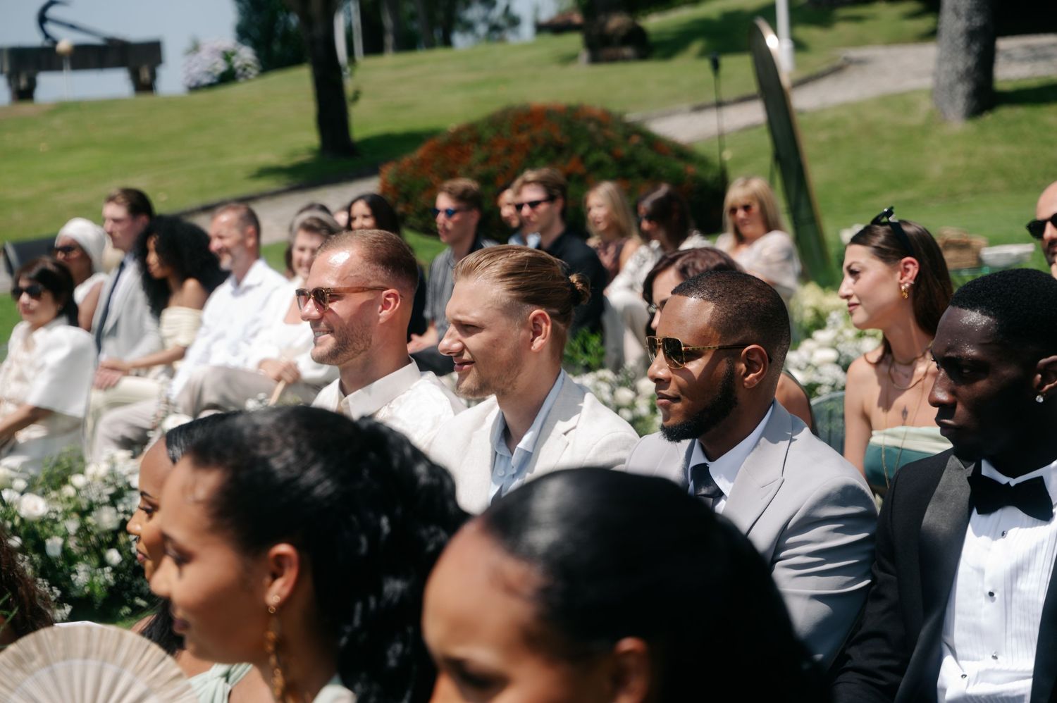 Wedding guests seated outdoors on white chairs during a sunny garden ceremony.