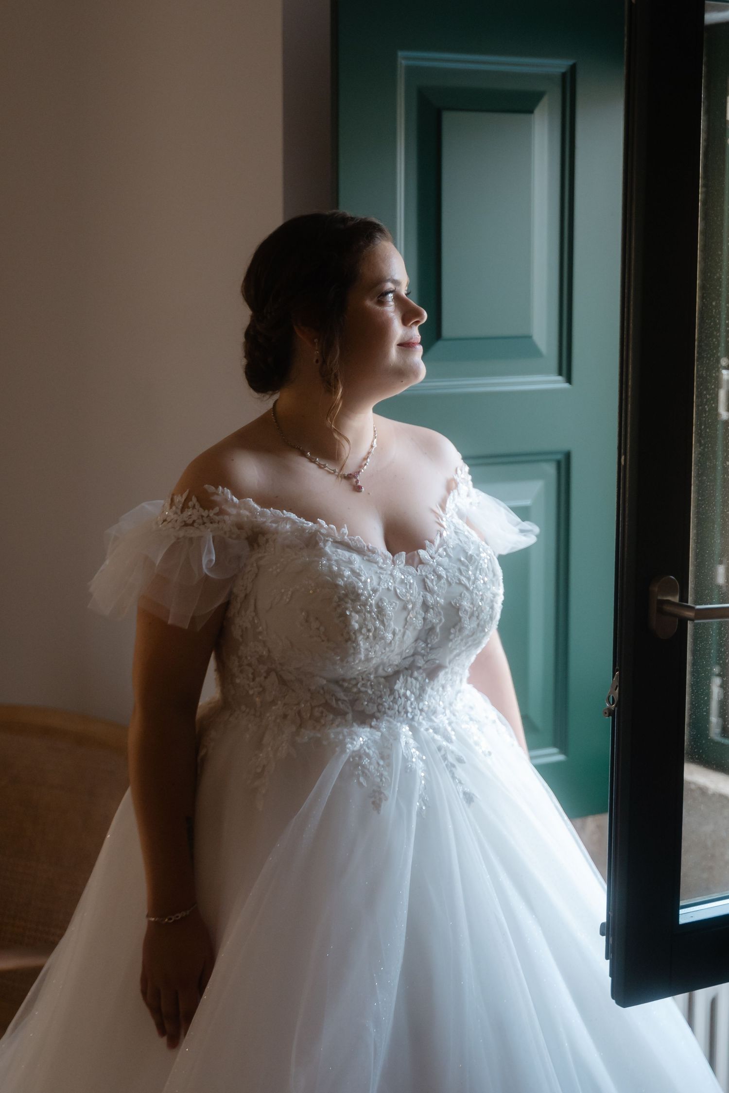 A bride in an elegant white lace wedding gown with cap sleeves poses near a green door in natural lighting.