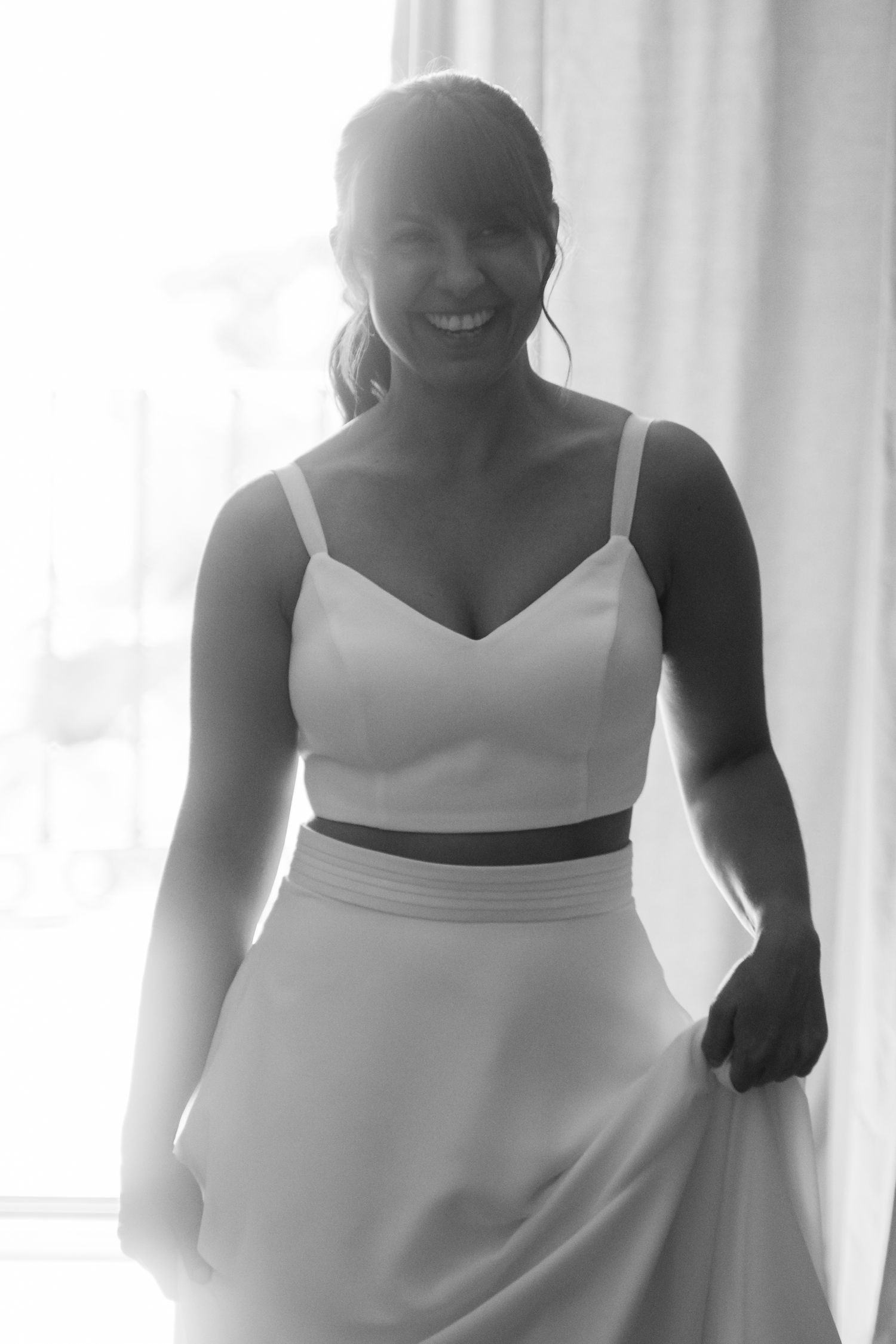 A person in a white formal dress with a fitted bodice and flowing skirt stands in front of a bright window.