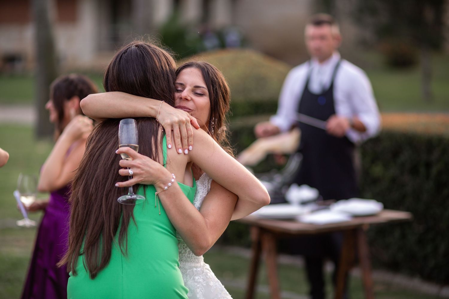 A heartfelt embrace at an outdoor wedding reception with a server in the background.