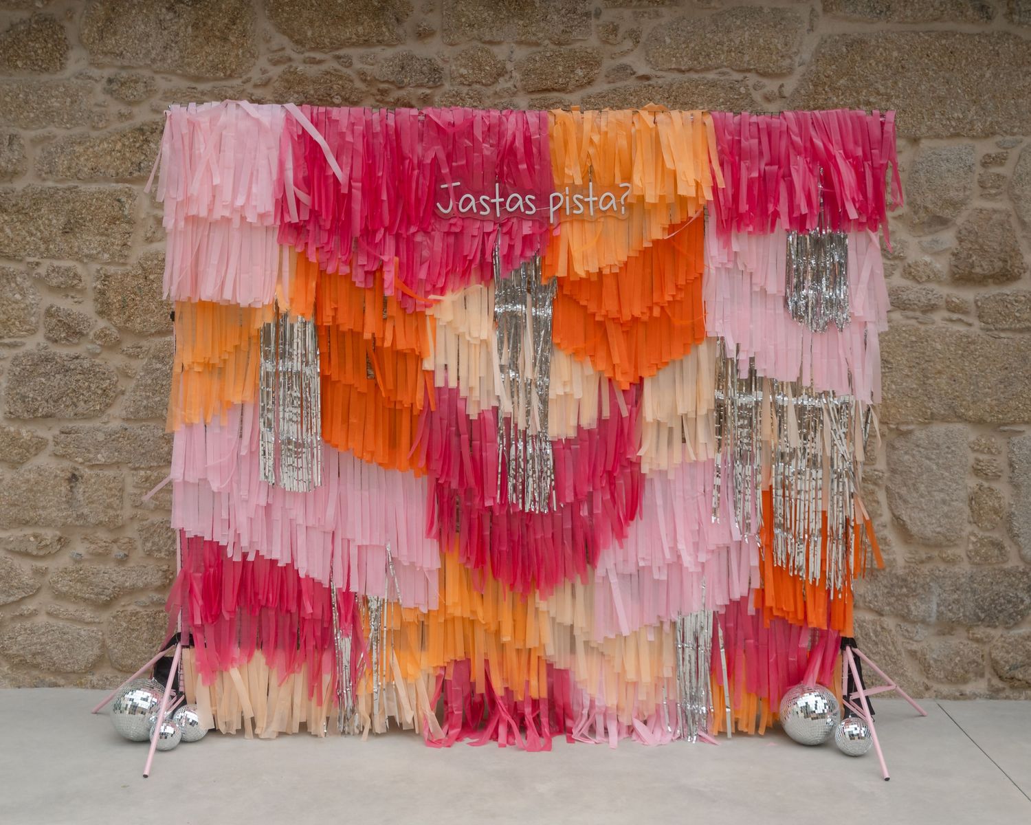 Colorful fringe streamers in pink, orange and coral hues hang against a rustic stone wall creating a festive backdrop.
