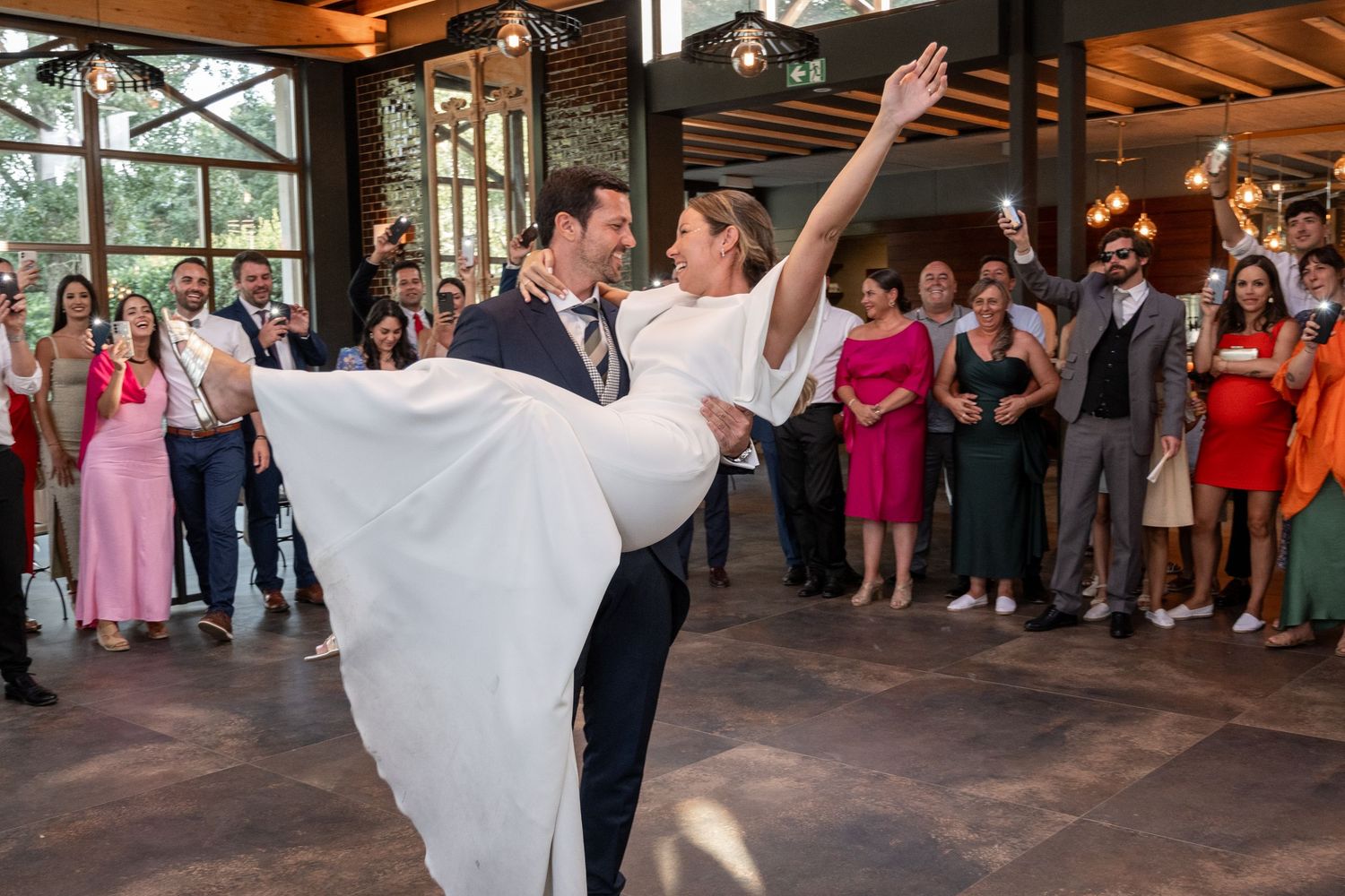 Wedding guests dance and celebrate on a rustic wooden dance floor at an indoor reception venue.