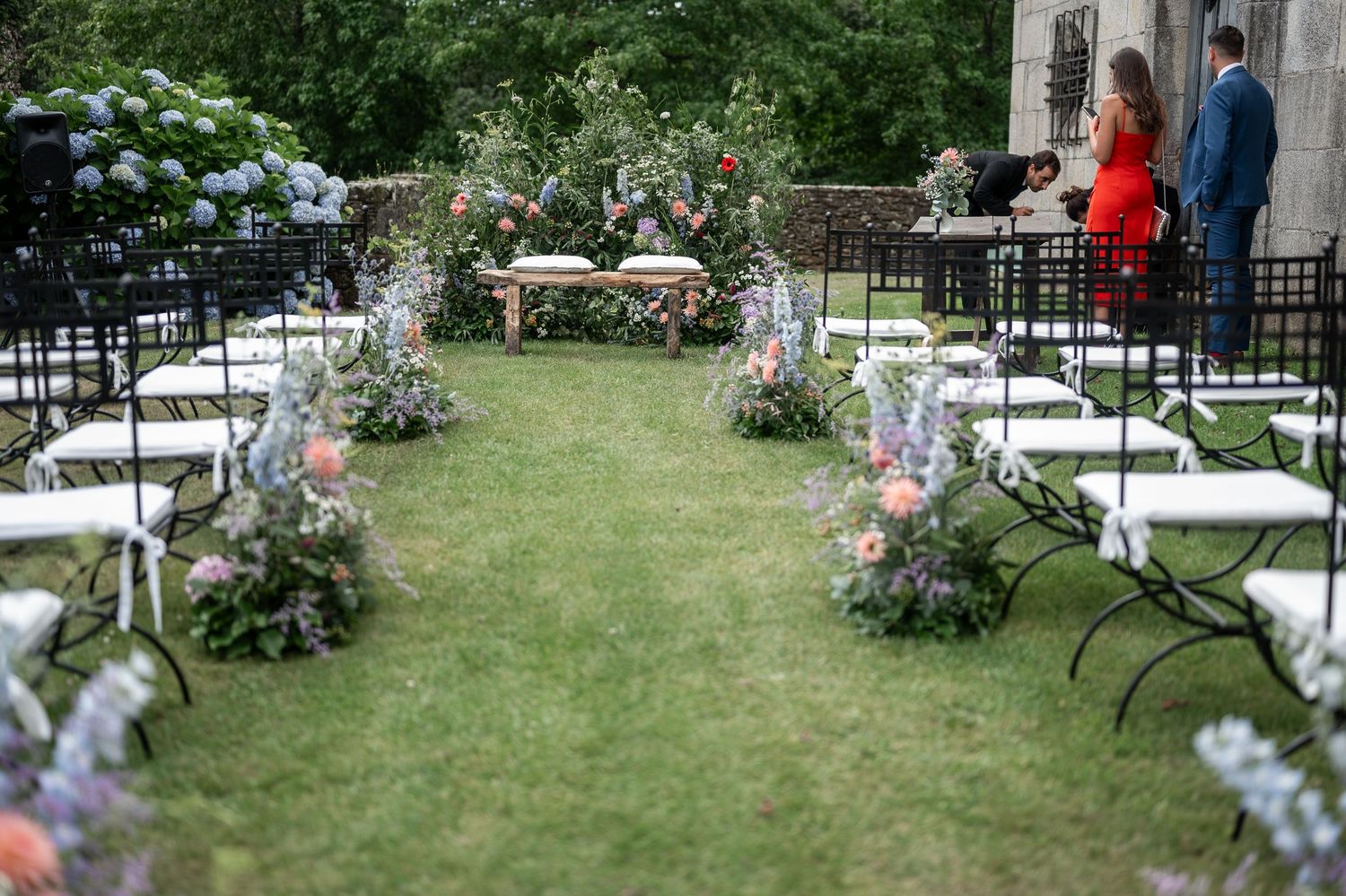 Outdoor garden wedding ceremony setup with white chairs and floral arrangements along a grassy aisle.
