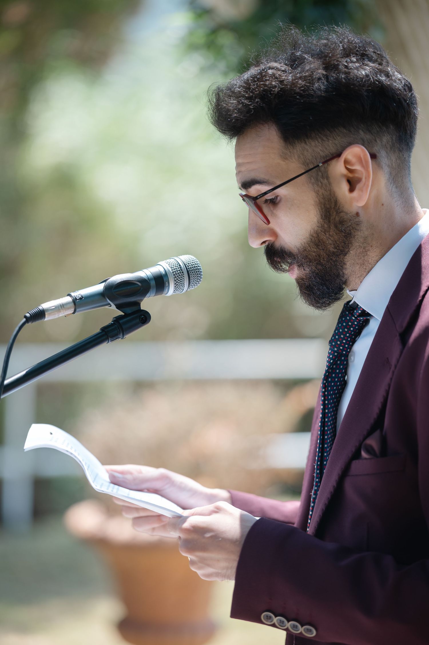 A speaker in a burgundy suit jacket reads from papers during an outdoor ceremony.