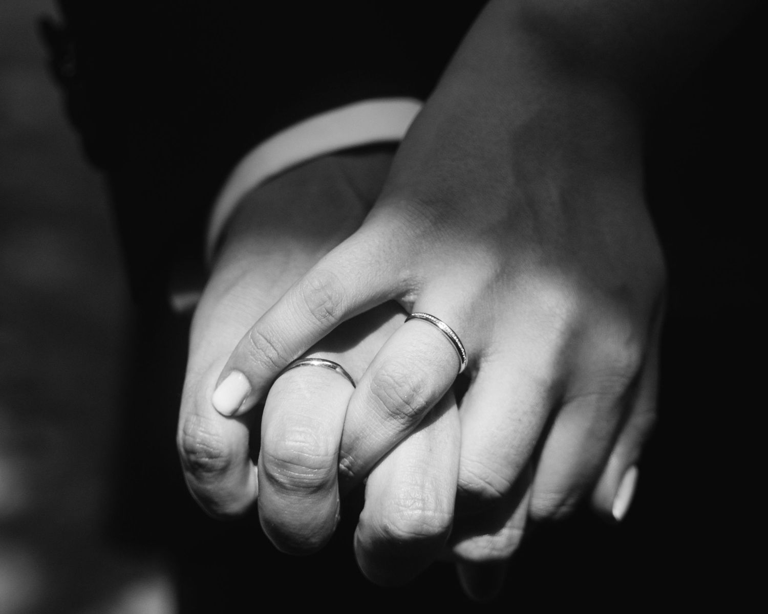 A black and white close-up photograph of hands wearing wedding rings against a dark background.