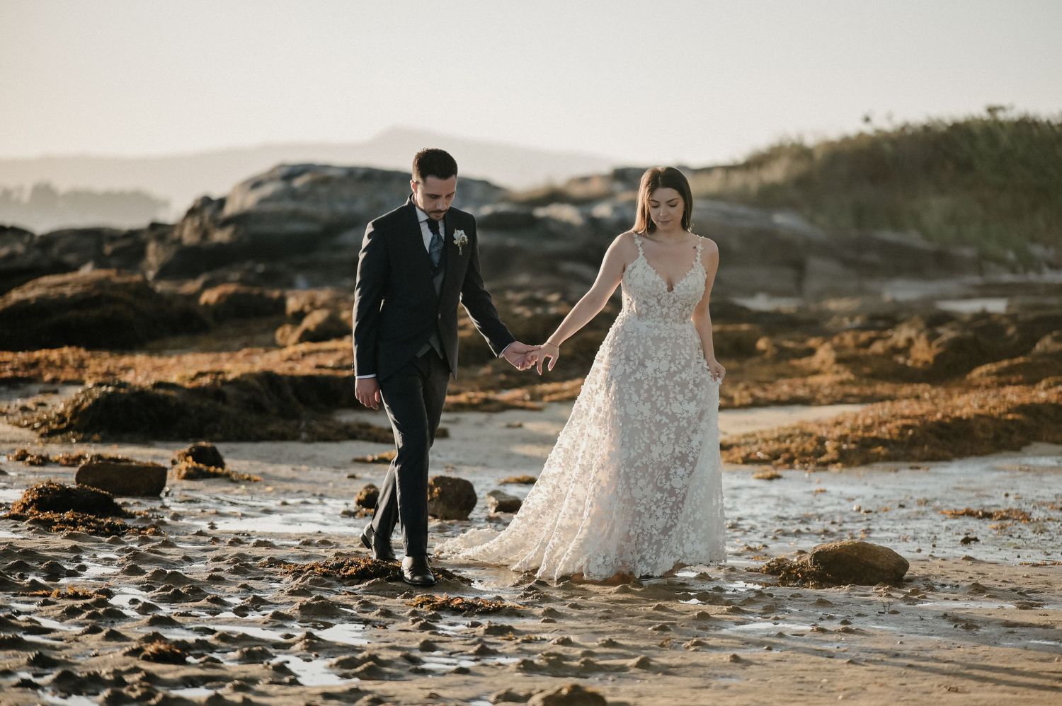 A couple walks along a rocky coastal beach at sunset in formal attire.