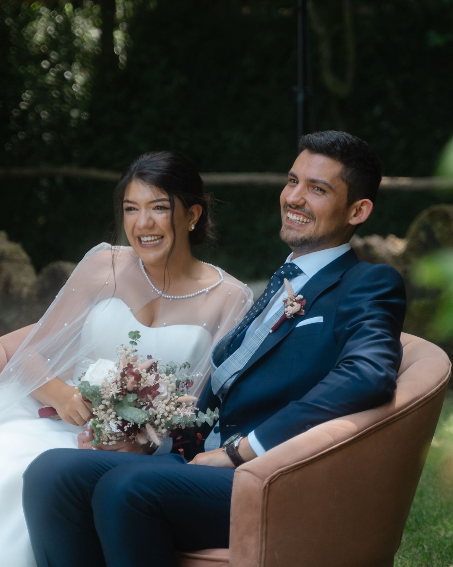 A bride and groom share a joyful moment while seated on a brown velvet chair in an outdoor garden setting.