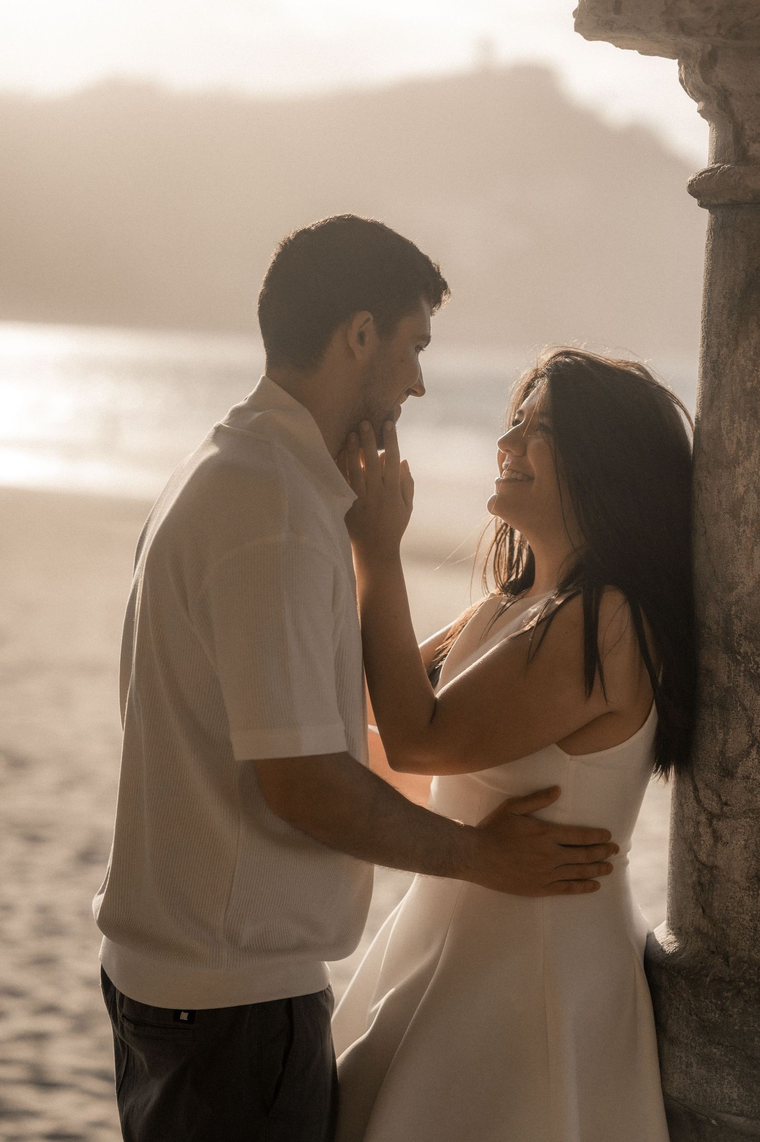 A romantic couple embraces by a pillar during sunset at the beach in a series of intimate poses.