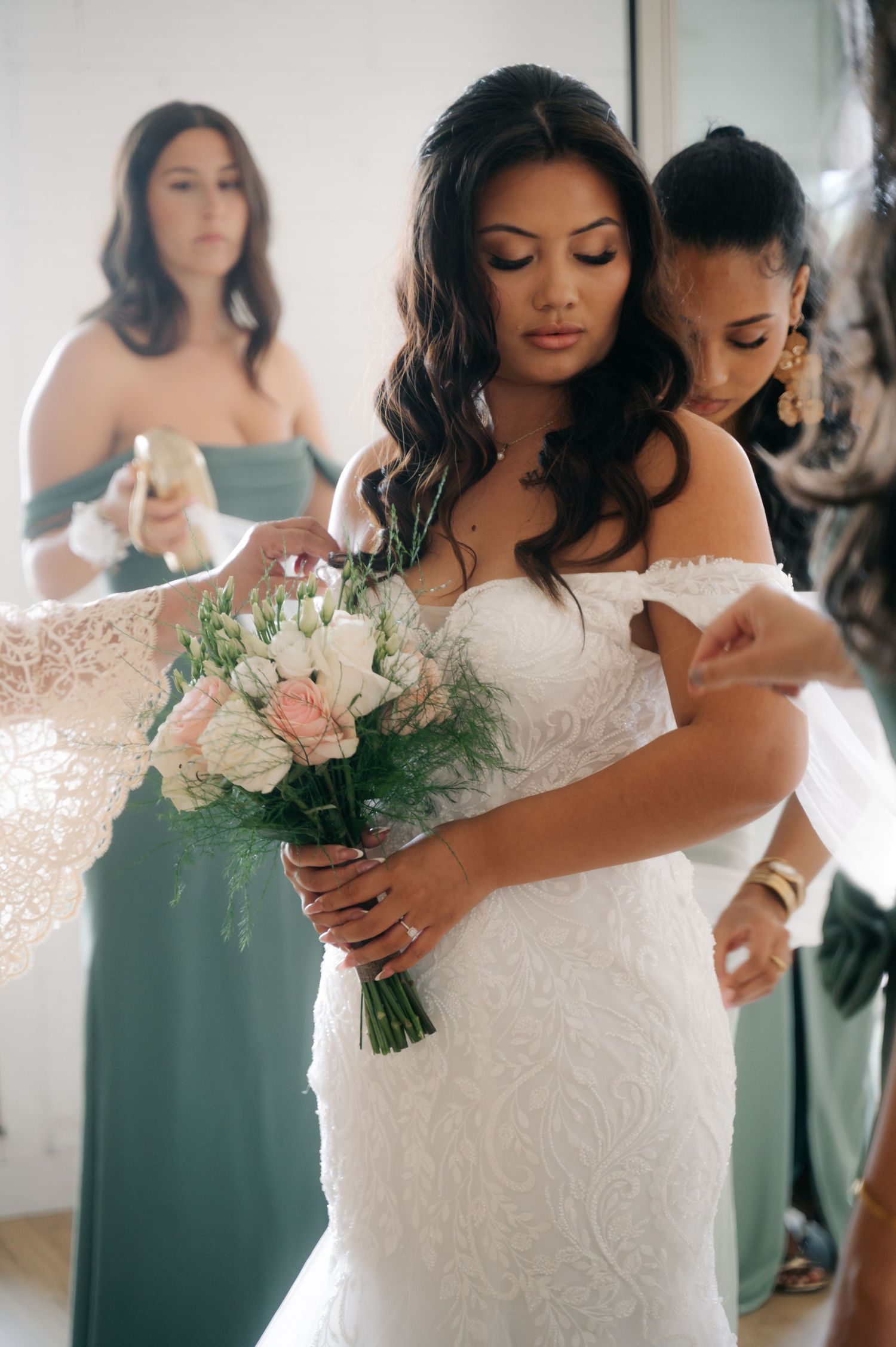 Bridesmaids in sage green dresses help adjust a bride's lace wedding gown in a getting ready sequence.