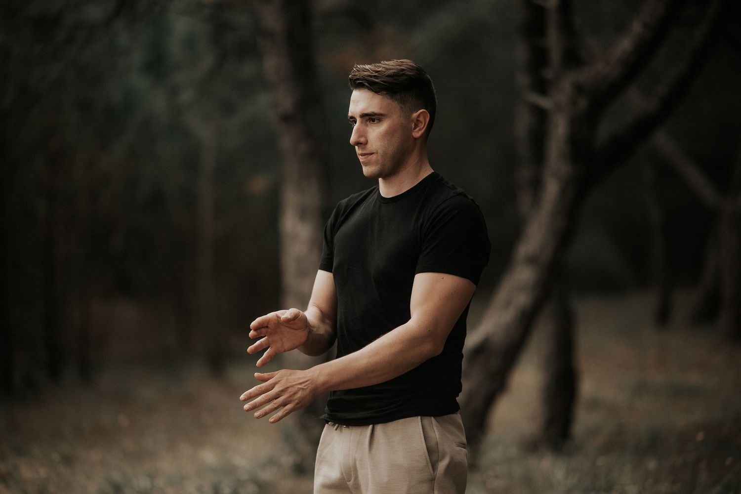 A person in a black t-shirt poses in a dark forest setting with dramatic lighting and shadows.
