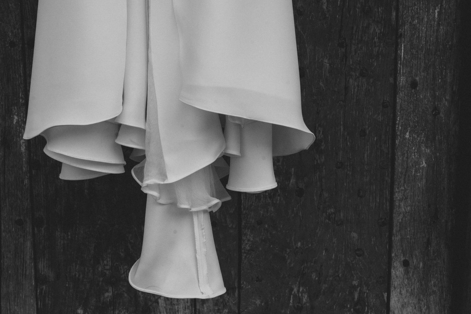Elegant white wedding dress with ruffled layers and flowing fabric hangs against a dark textured wall.