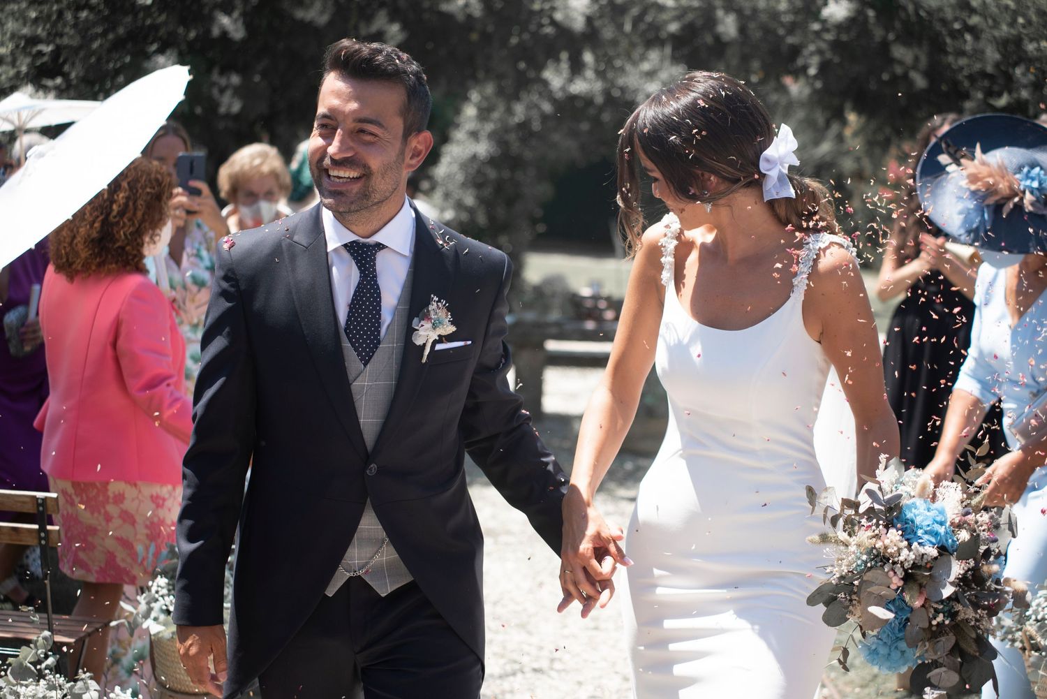 Wedding party celebrates with confetti toss outdoors on sunny day.
