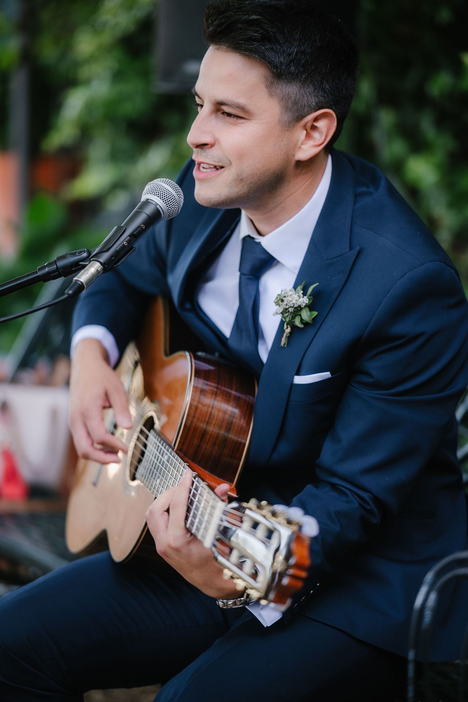 A musician in a navy suit plays acoustic guitar during an outdoor wedding performance.