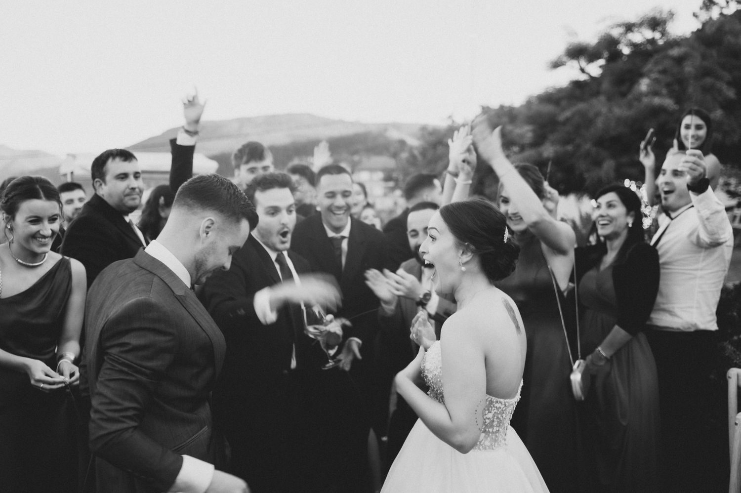 Wedding party and guests dance enthusiastically outdoors with raised arms against a scenic mountain backdrop.