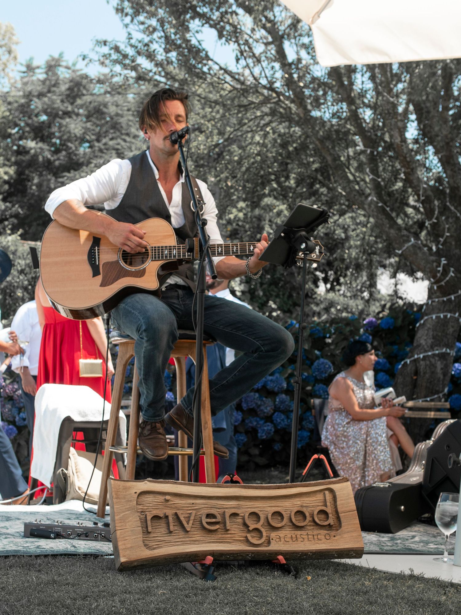 Musicians perform outdoors with acoustic guitars and wooden stools at an outdoor gathering.