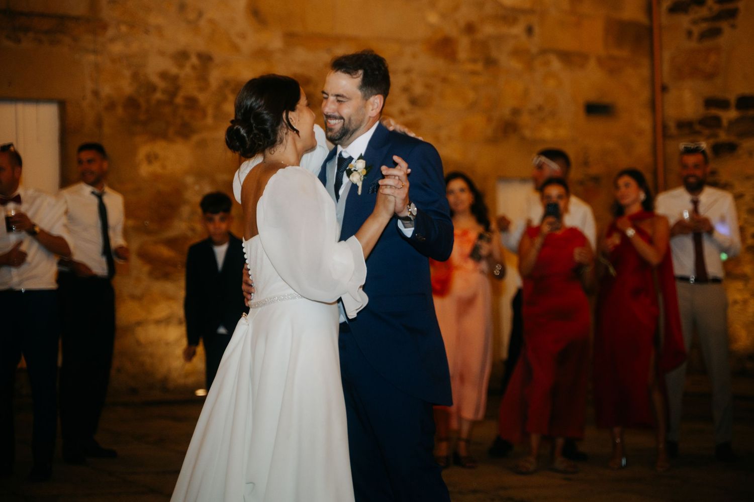 A couple shares their first dance at a wedding reception with bridesmaids in red dresses watching in the background.