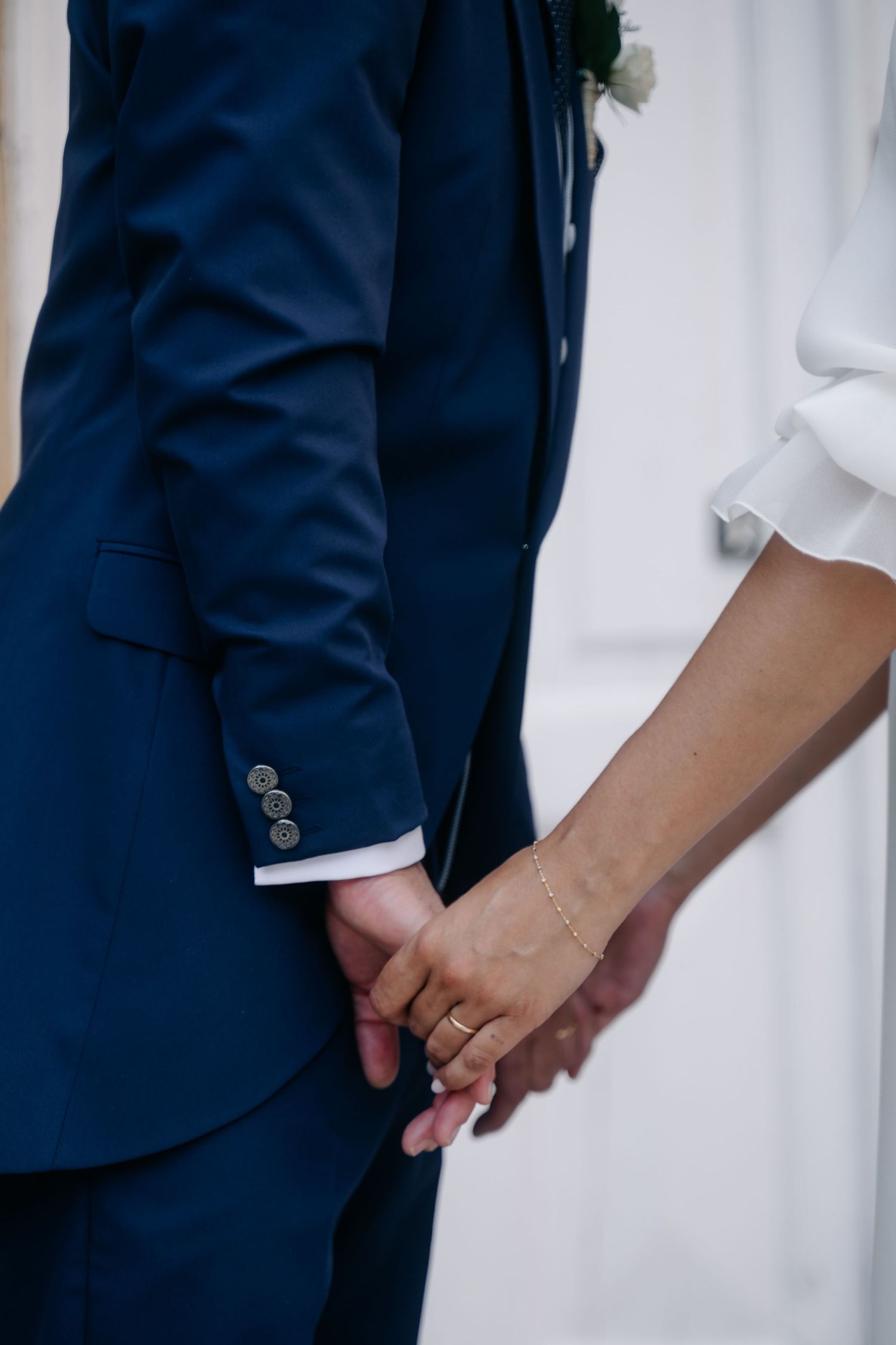 Close-up detail of hands wearing wedding rings against navy suit and white dress fabrics.