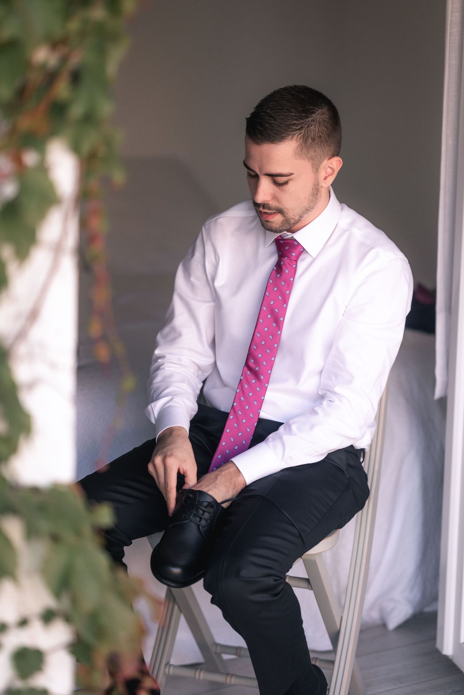 Professional dressed in crisp white shirt and pink tie adjusts his shoe while seated on a white chair.