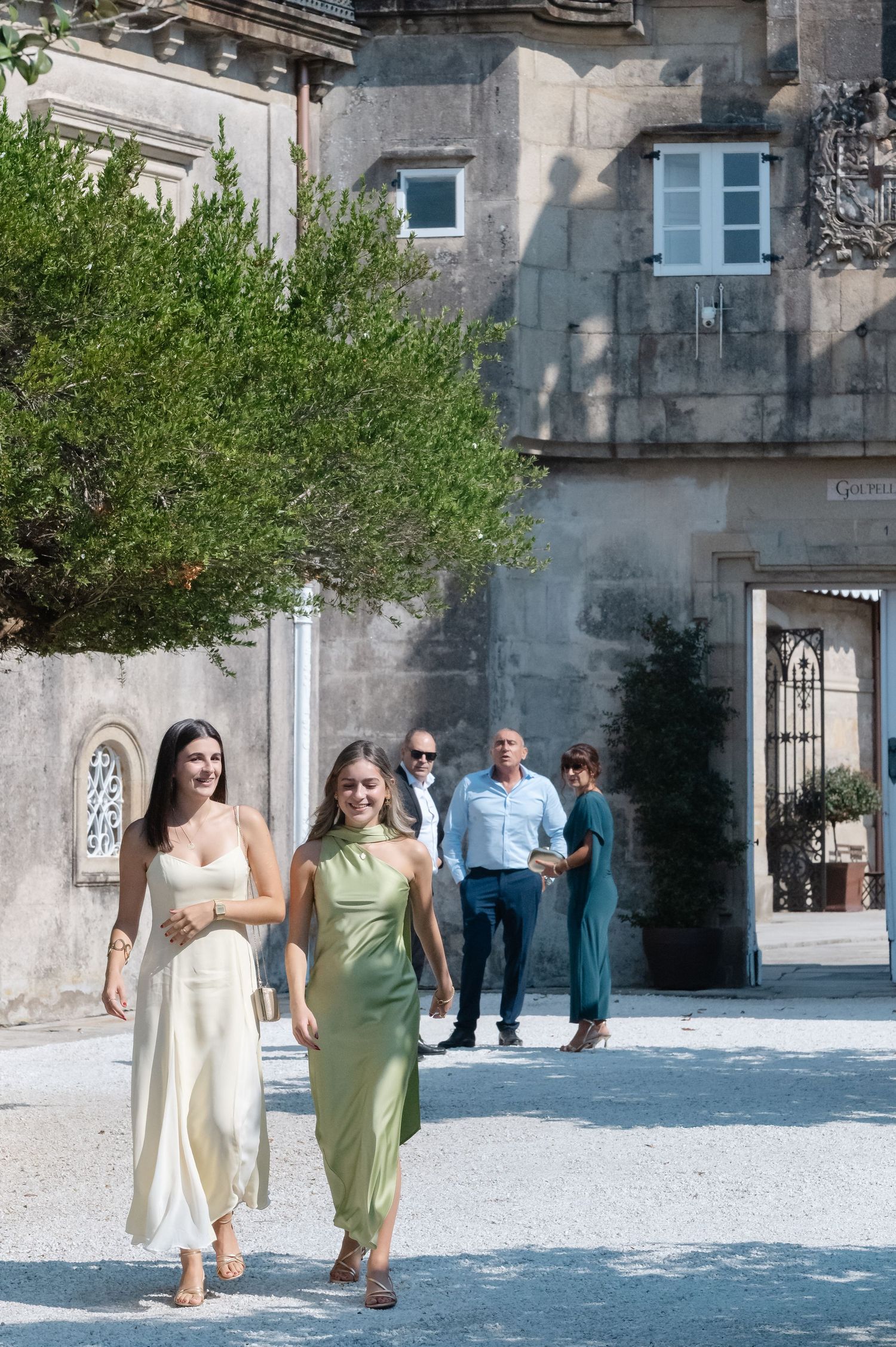 Wedding guests in pastel dresses walking across a stone courtyard of a historic building.