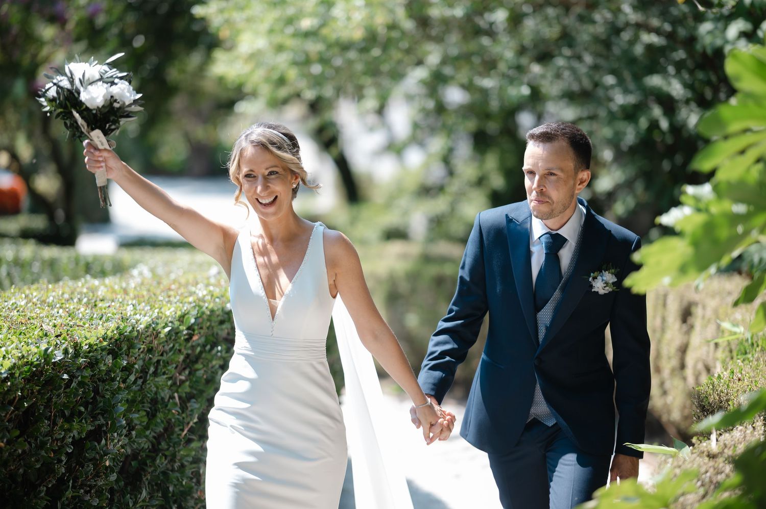 A couple walks and laughs together outdoors in formal wedding attire on a sunny day.