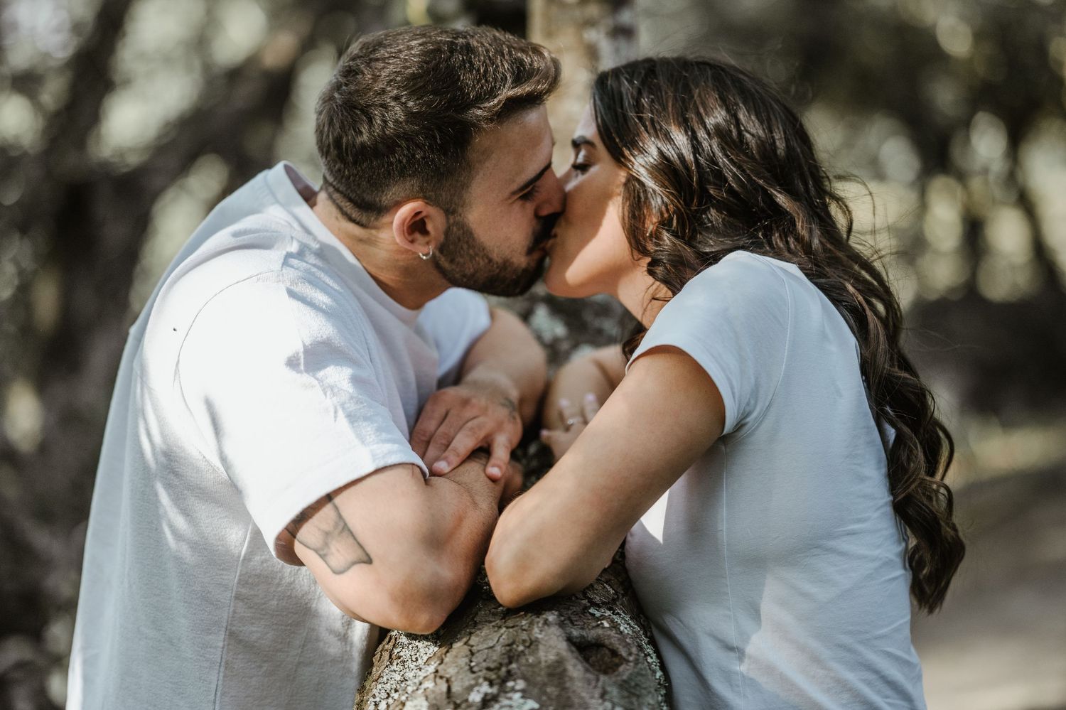A romantic couple shares a tender kiss outdoors in natural lighting with trees in the background.