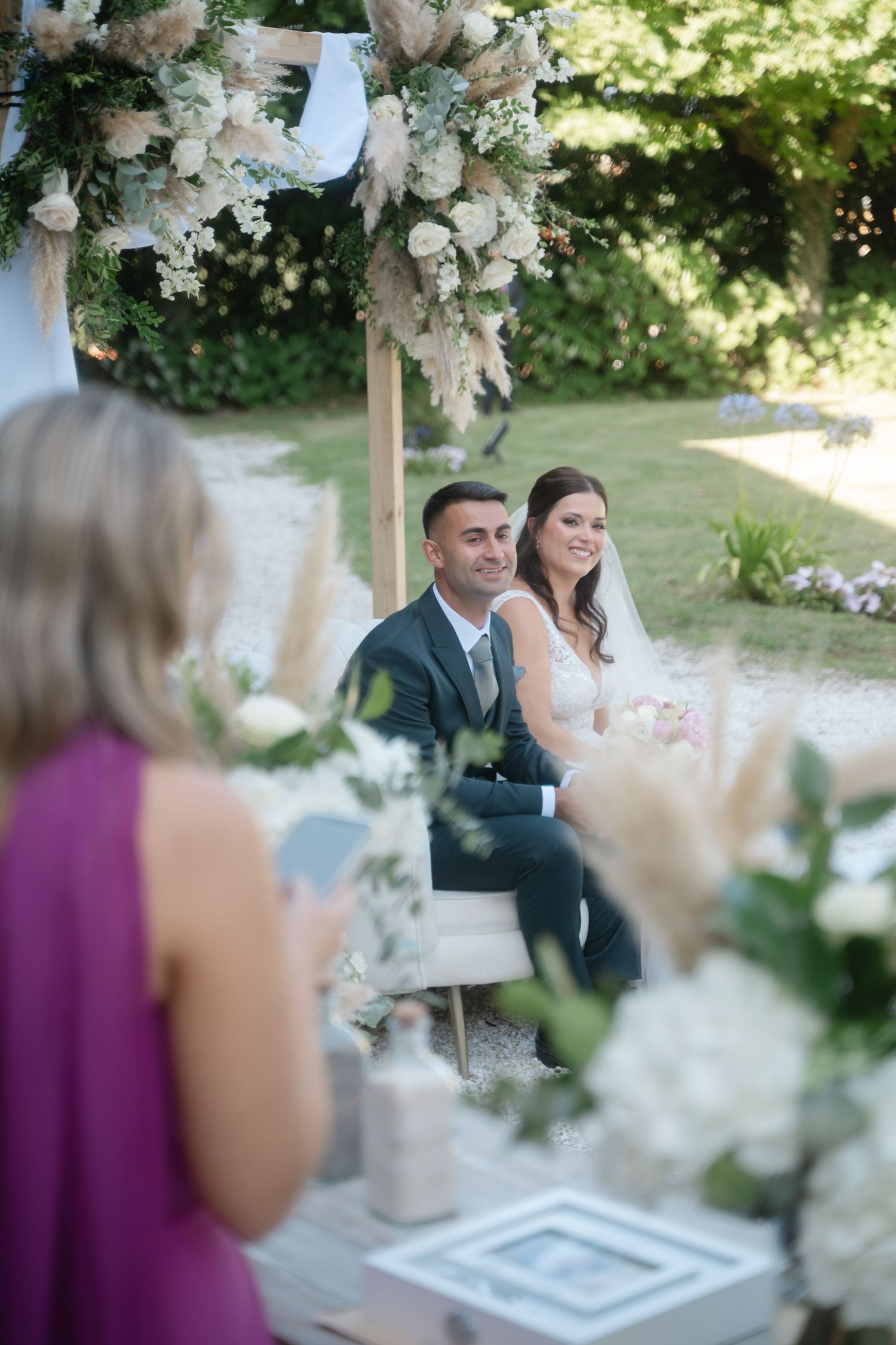 Outdoor wedding ceremony setup with white floral arch and guests seated in garden setting.