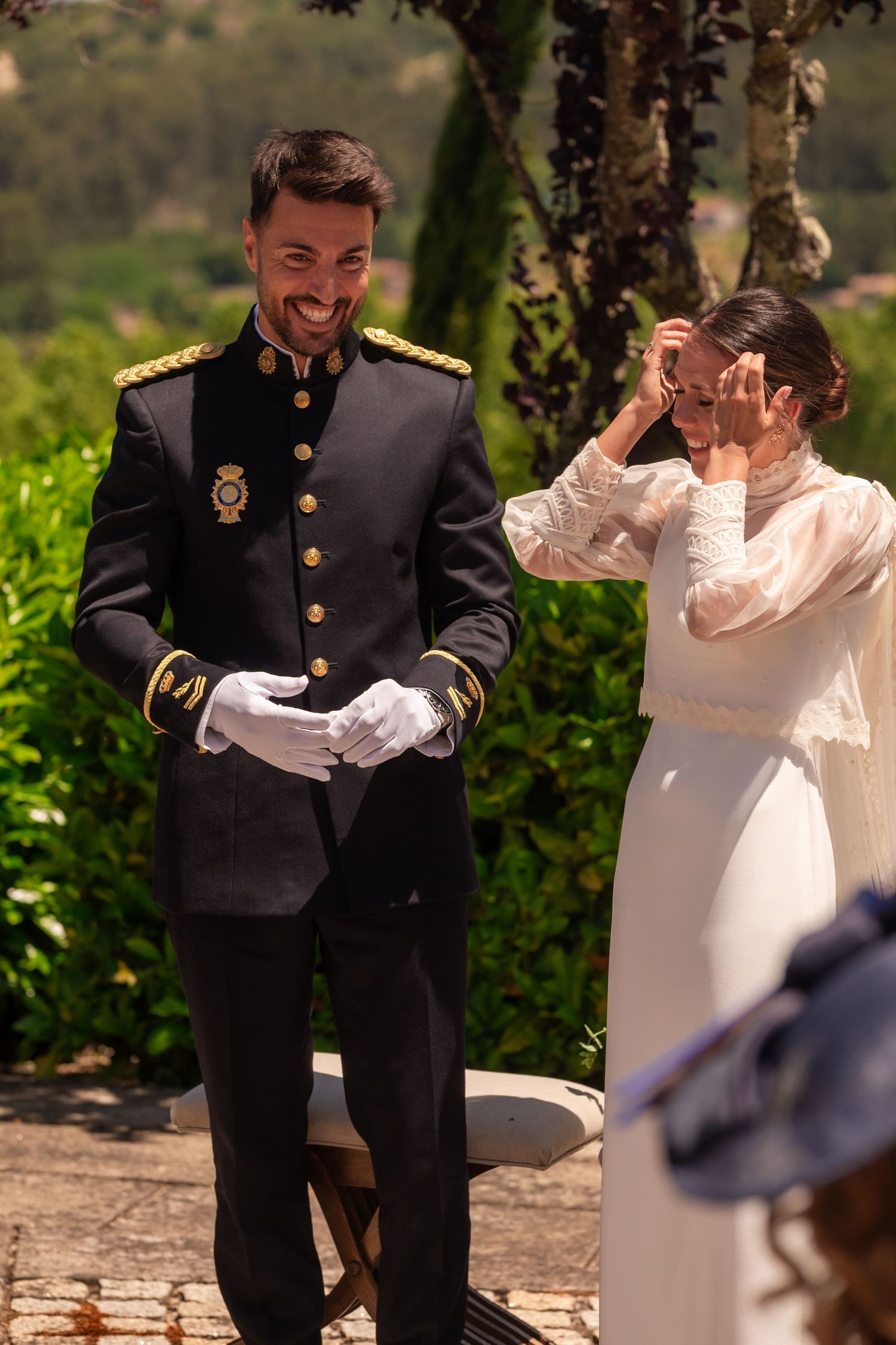 Couple celebrating during outdoor ceremony with formal military attire against garden backdrop.