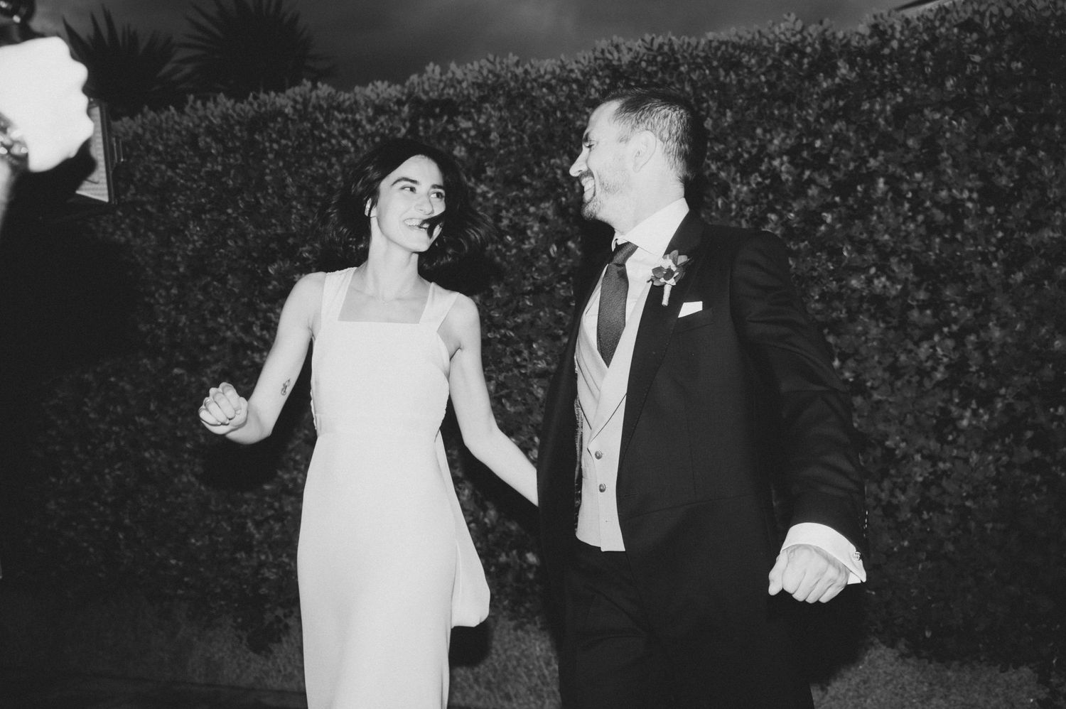 A black and white photo of newlyweds walking hand in hand beside a garden hedge.