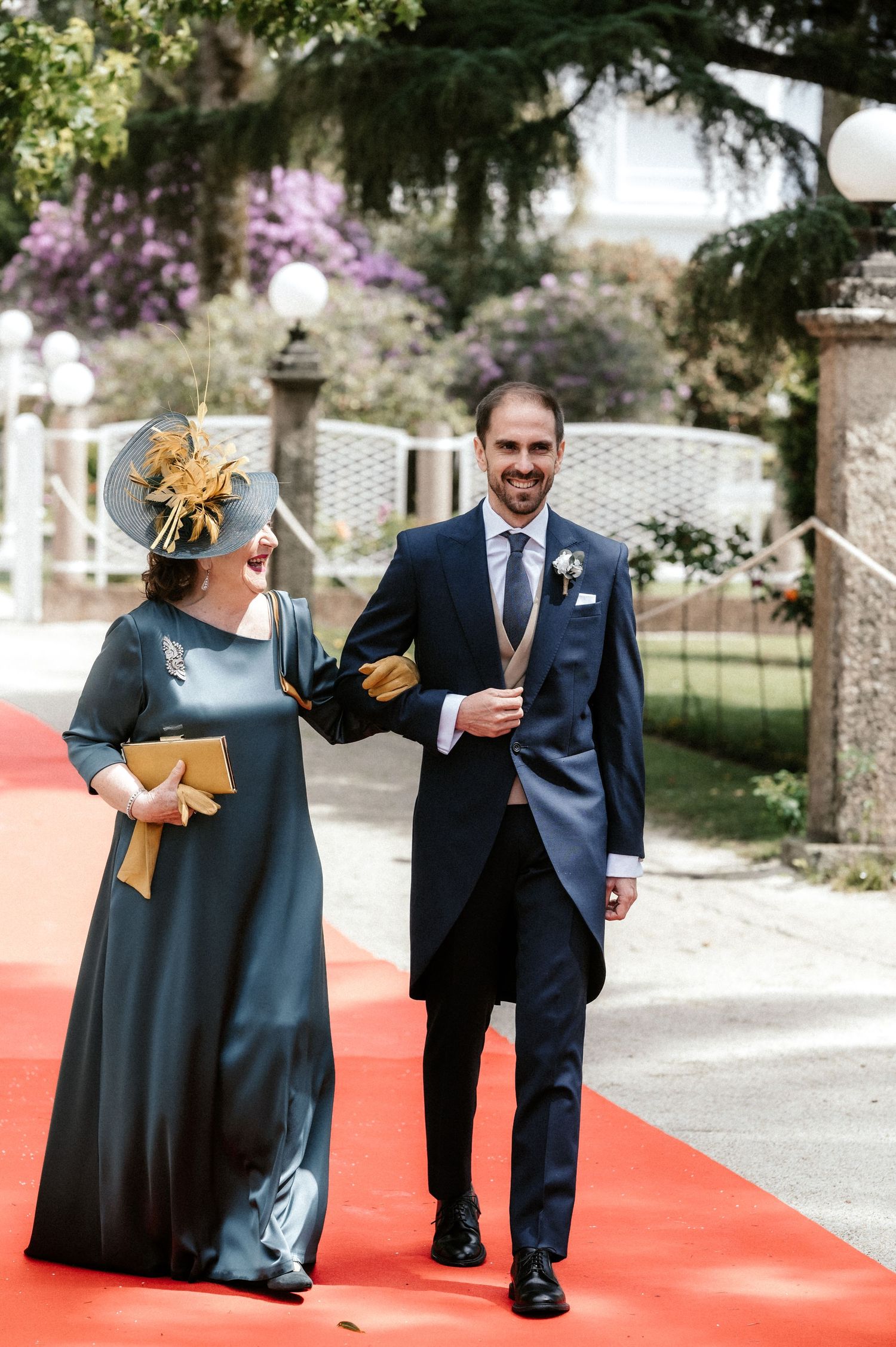Guests in formal attire walk along a red carpet path in a garden setting during a wedding celebration.