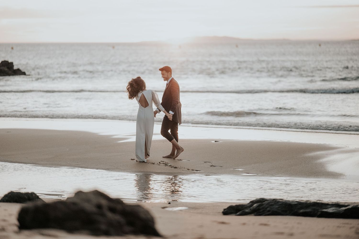 A couple walks along a misty beach shoreline during sunset while waves crash against the sand.