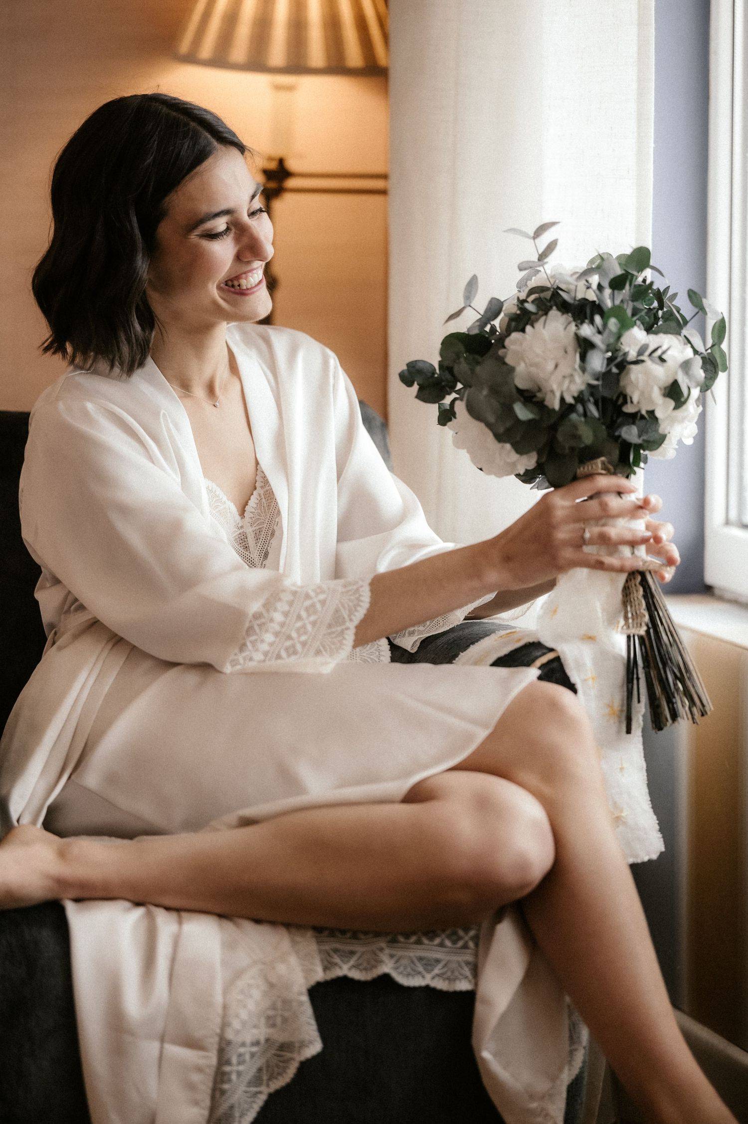 A figure in a white robe sits by a window holding a bouquet of flowers in a series of romantic boudoir photos.