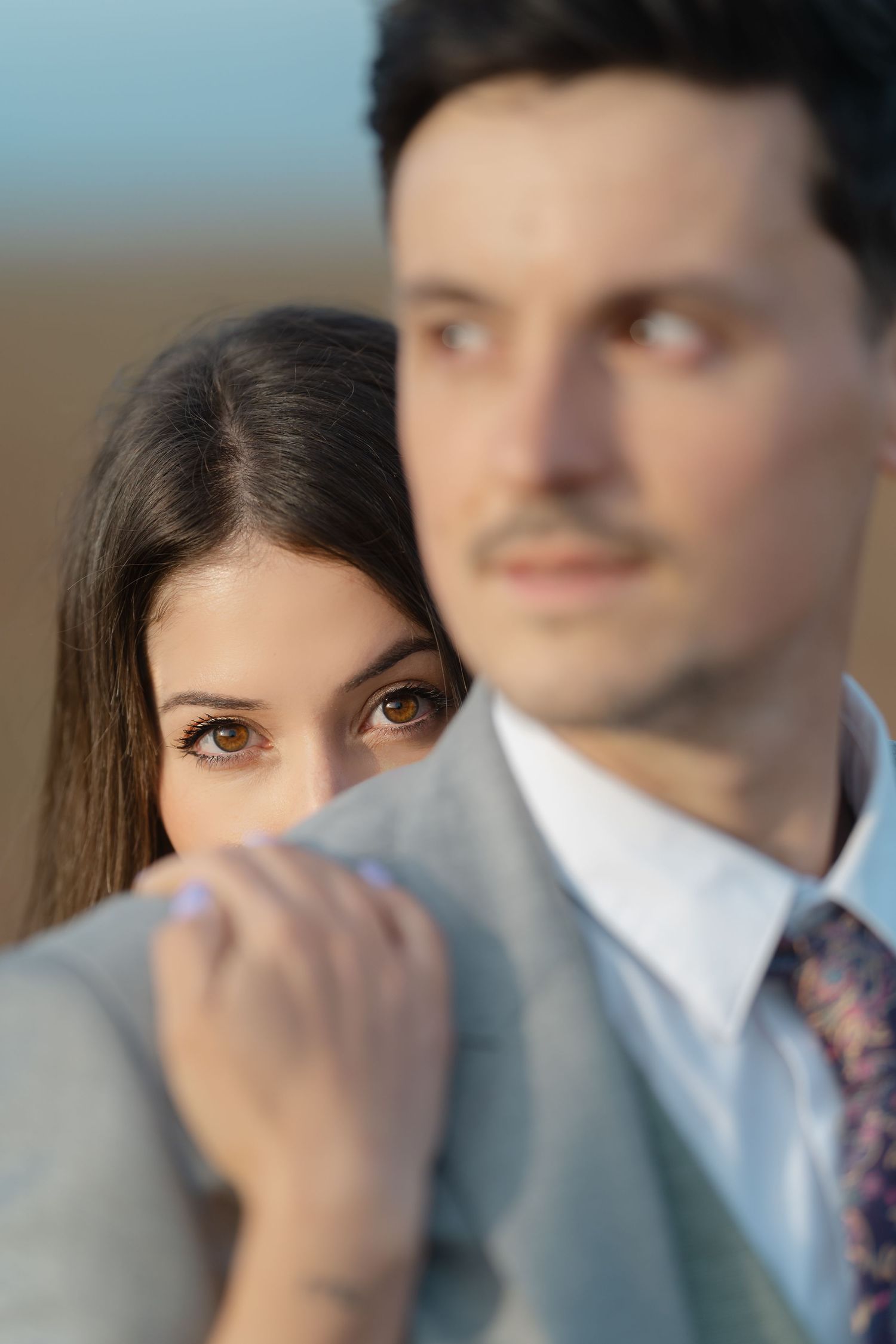A woman peeks over a man's shoulder as he gazes away, both dressed elegantly in soft natural light.