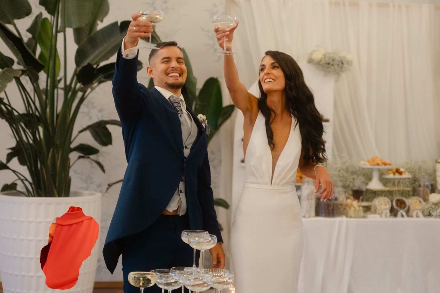 A couple in wedding attire raise their glasses in celebration at an elegant indoor reception with white decor and plants.